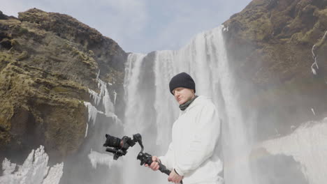 videographer holding camera on gimbal under waterfall in landscape of iceland on sunny winter day