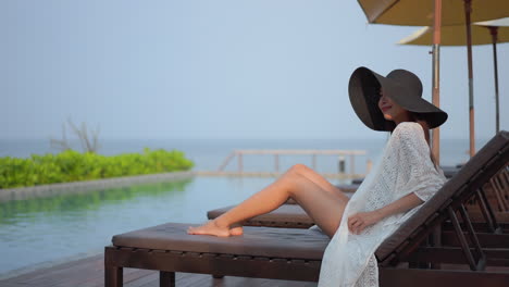 asian woman with a swimsuit and white lace cover up sits on a wooden sun lounger by the poolside