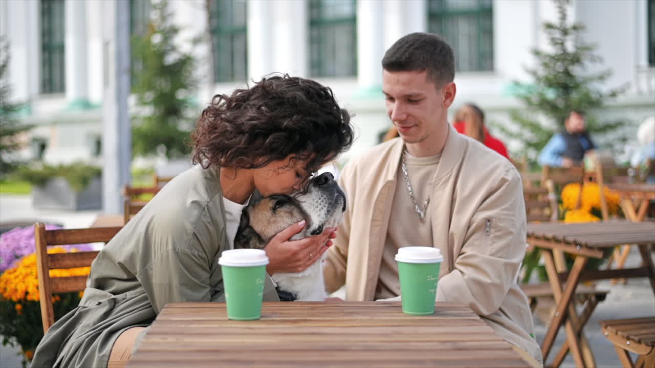 A happy couple outdoors near a cafe. Petting their dog, coffee on the table. Autumn atmosphere. Slow motion