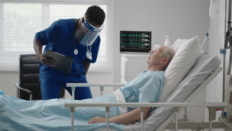 an elderly male patient lying in a hospital bed connected to an ecg machine talks to a black doctor in a protective mask