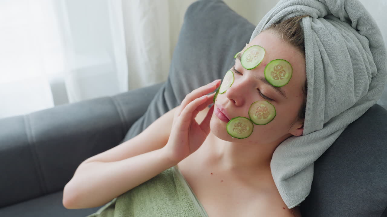 Woman seated on couch wrapped in towel placing cucumber slices on face during skincare routine, relaxing with eyes closed in bright room, serene expression, calm beauty ritual promoting glow