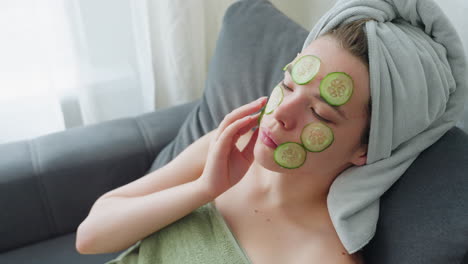 woman seated on couch wrapped in towel placing cucumber slices on face during skincare routine, relaxing with eyes closed in bright room, serene expression, calm beauty ritual promoting glow