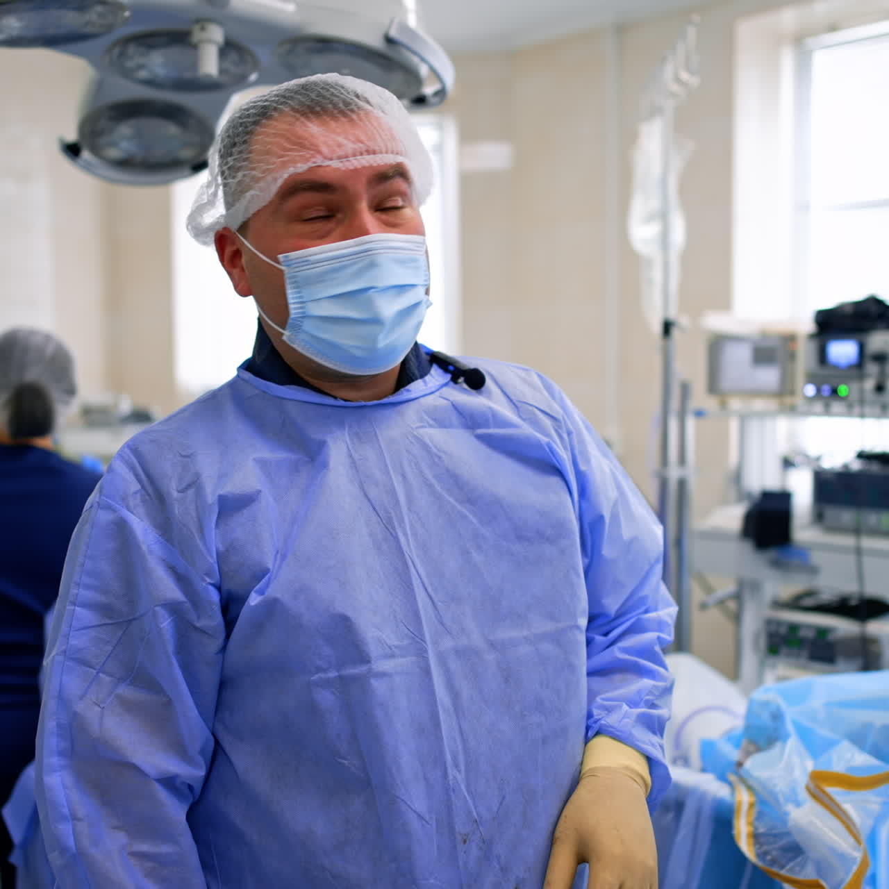 Positive male surgeon wearing mask talking to camera. Nurses at backdrop work with tools and instruments.