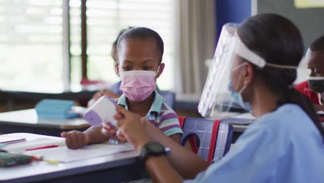 Diverse-medical-worker-showing-schoolgirl-how-to-measure-temperature,-all-wearing-face-masks,