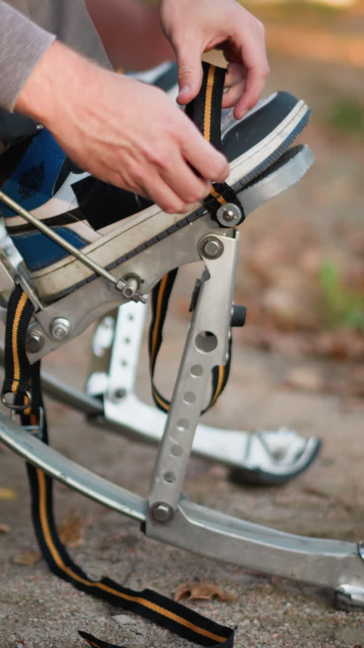Close-up of person adjusting spring stilts on gravel path with fallen leaves, wearing gray sweatpants and dark sneakers, ground-level focus on preparation and equipment setup outdoors
