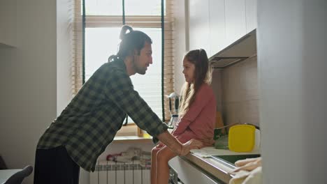 un hombre moreno con una camisa a cuadros verdes se apoya en la mesa y se comunica con su pequeña hija morena en un vestido rosa en una cocina moderna