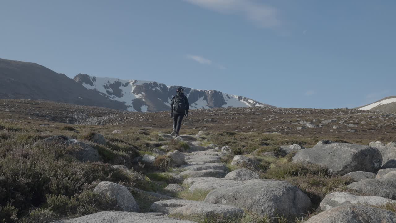 plano general de una mujer caminando hacia el pico nevado de la montaña escocesa con un sendero rocoso como líneas principales