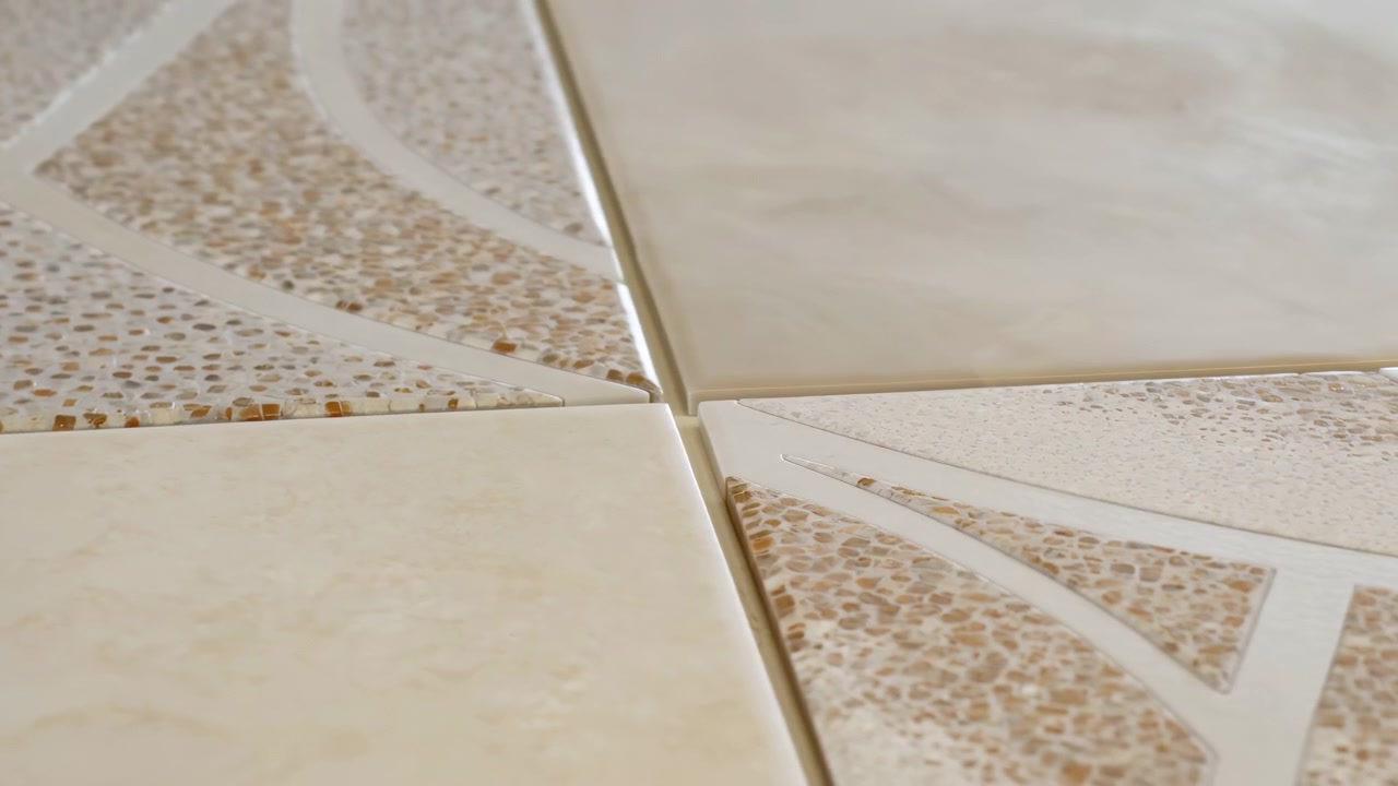 Low oblique close-up of four ceramic or stone tiles meeting at central grout junction, showing smooth cream slabs and textured pebble-pattern tiles with beige and light-brown speckles