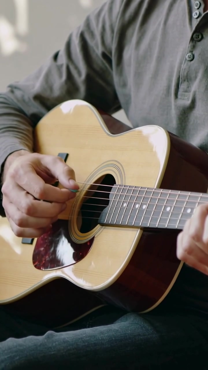 Video verticale: Uomo in maglietta henley grigia che porta la chitarra in grembo e strimpella con il plettro a casa per fare pratica.