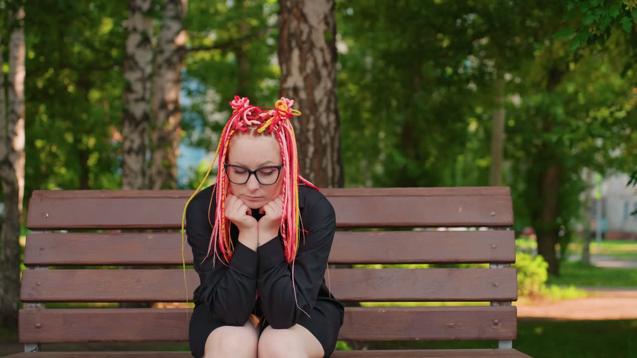 화려한 머리를 한 고요한 여성, 평화롭게 야외에 앉아 있는 생동감 넘치는 드레드락을 한 생각에 잠긴 여성, 잎이 무성한 나무 아래 나무 벤치에 앉아 있는 네온 색상의 드레드락을 한 사색에 잠긴 여성
