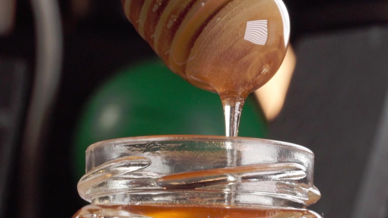 Miele che cola da un mestolo di legno in un barattolo di vetro aperto, mostrando un liquido dorato lucido, una consistenza viscosa e un flusso lento su sfondo scuro.