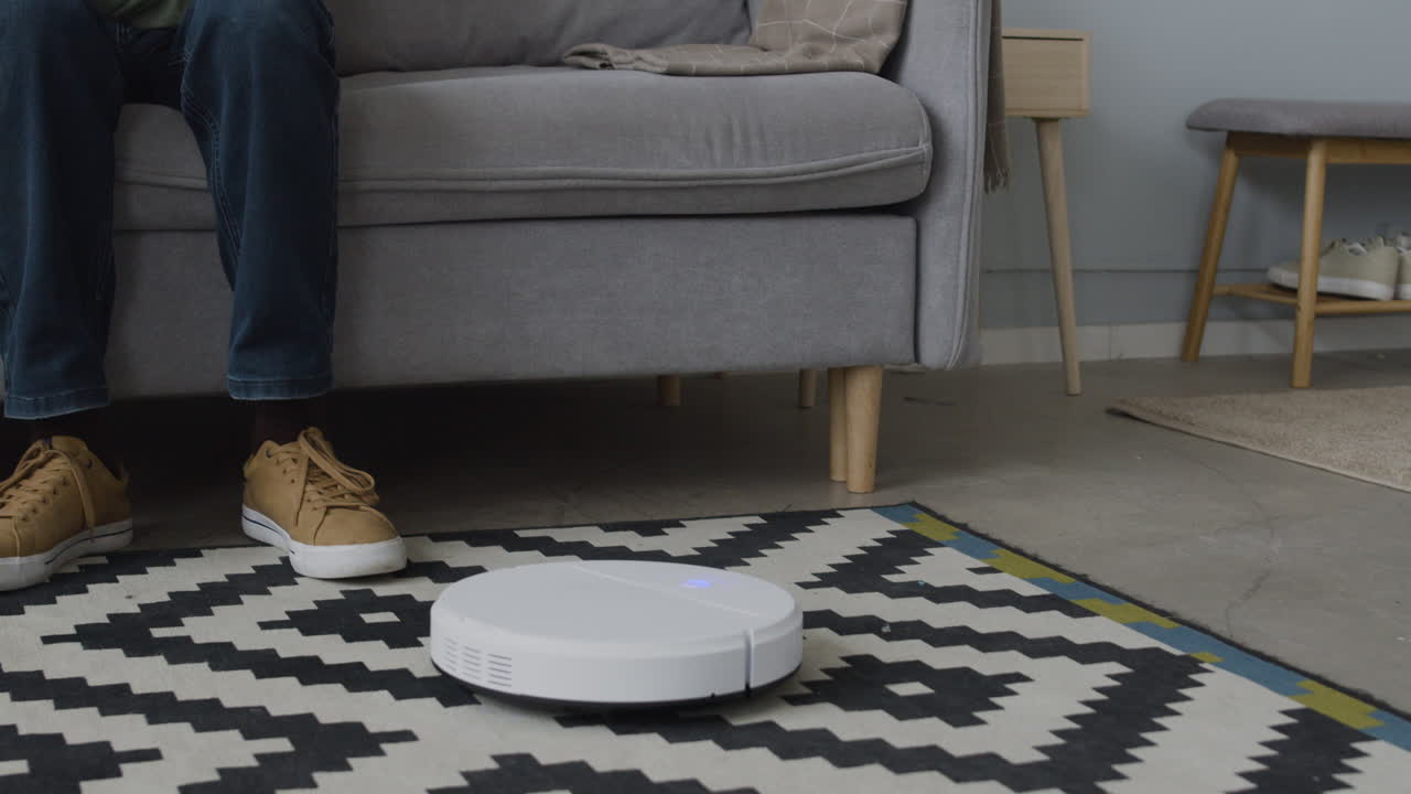 неузнаваемая пара отдыхает на диване и поднимает ноги, пока автоматический пылесос убирает комнату