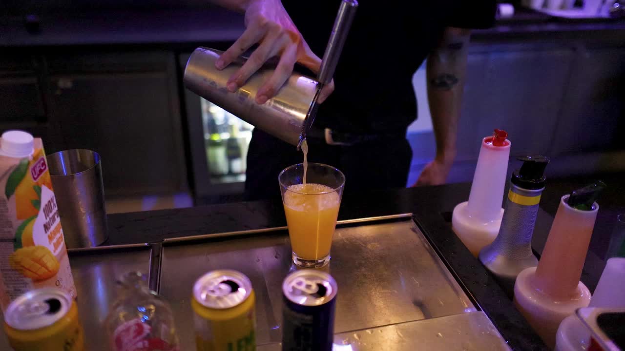 Bartender pours vibrant cocktail over ice in dimly lit bar with colorful ambient lighting