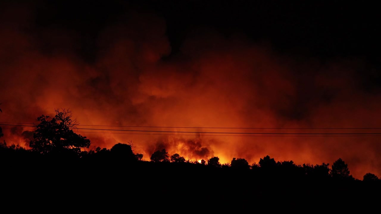 Scatto drammatico che cattura un intenso incendio boschivo che circonda un villaggio rurale portoghese di notte, con fiamme altissime, fumo denso e cieli incandescenti durante un'ondata di calore record europea legata a El Niño.
