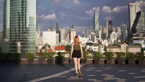 Una-Hermosa-Joven-Camina-Lentamente-Sobre-Una-Azotea,-Admirando-La-Vista-Panorámica-Del-Horizonte-De-Bangkok