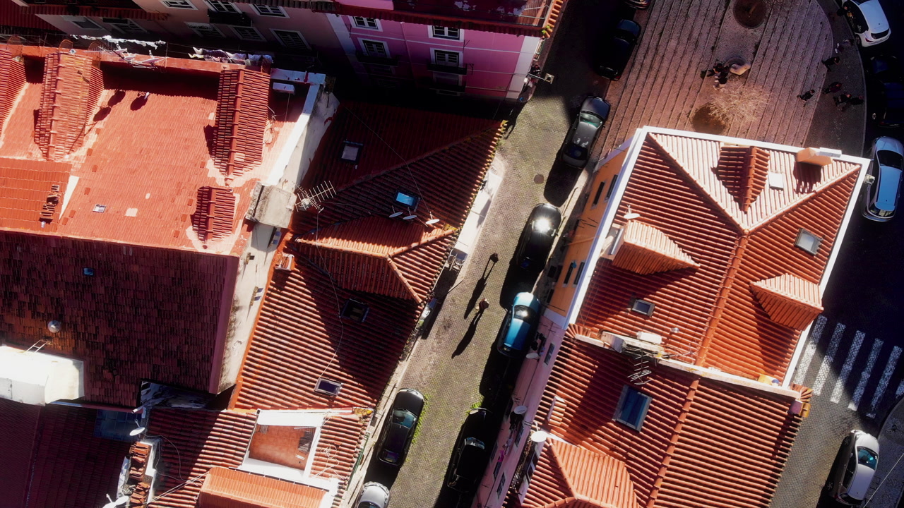 Spinning drone descending over Lisbon street with pedestrians, cars and red rooftops in sunlight