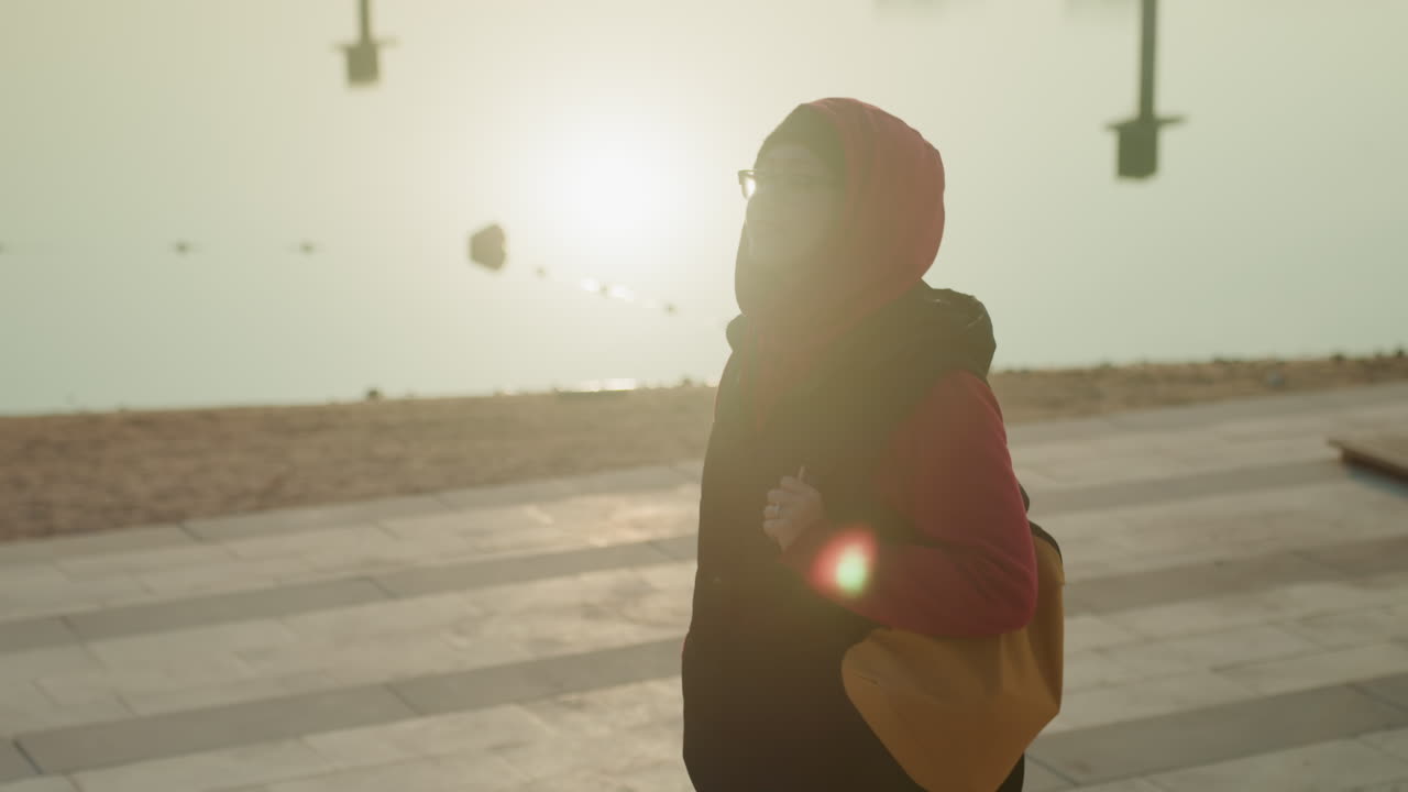 Wanita Arab di tepi pantai berjalan saat matahari terbit, cahaya latar keemasan dengan burung-burung di atas kepala, tas kuning dan kacamata hitam, jalan setapak beraspal dan cakrawala laut, angin sepoi-sepoi, langkah optimis, efek suar lensa sinematik