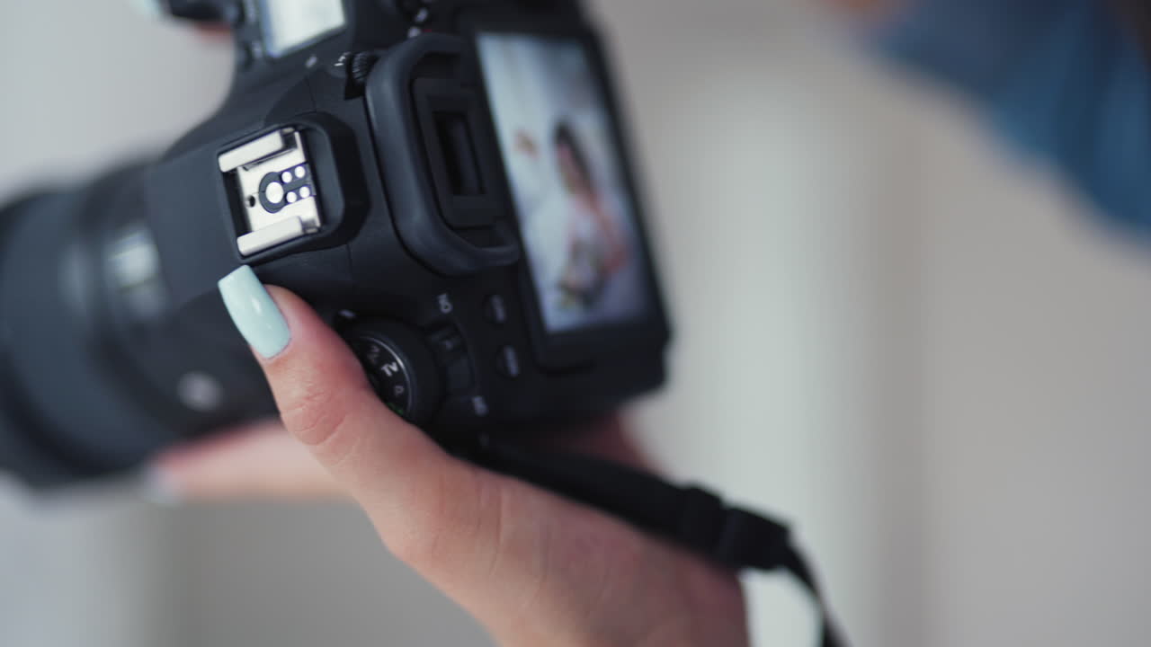 Female photographer with long dark hair is shooting by camera a pretty bride. Attractive young bride with flowers posing to the professional photographer on a white bed in studio.