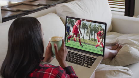 compuesto de una mujer sentada en casa sosteniendo un café viendo un partido de hockey en una computadora portátil