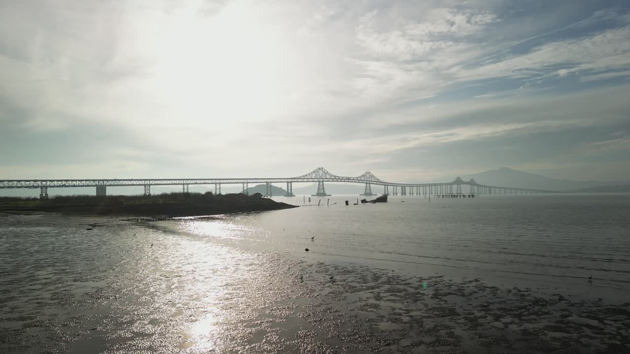 A low angle aerial view showcasing the Richmond–San Rafael Bridge with calm waters and the shore of Point Molate.