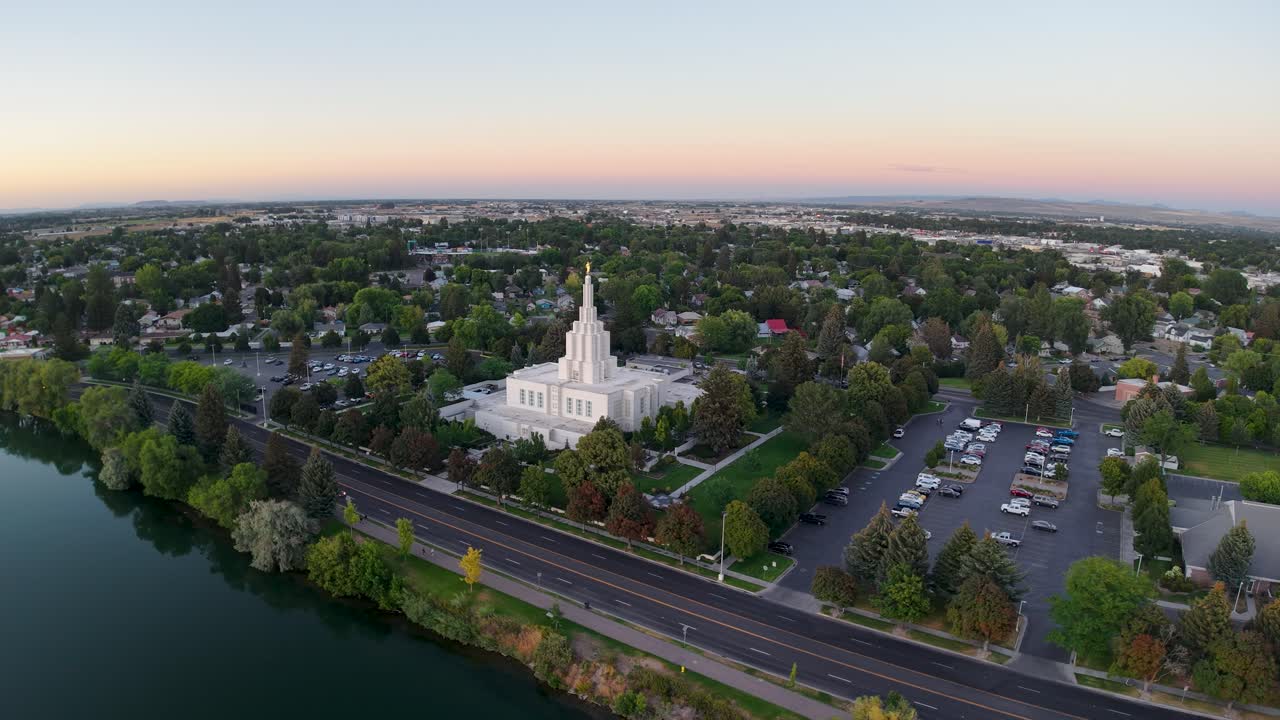 Dronebilde i luftperspektiv av Idaho Falls-tempelet og det omkringliggende bybildet langs Snake River med trær, veier og biler under solnedgangen