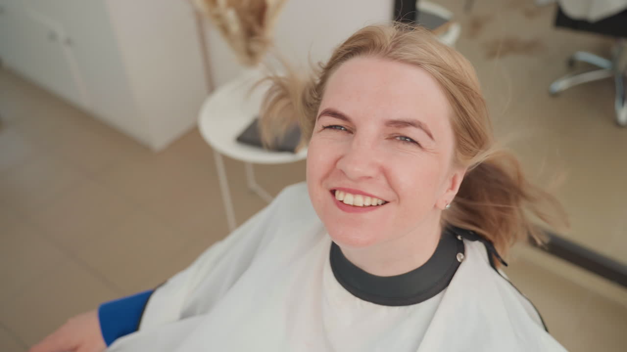 Vaalea nainen kampaamossa hymyilee lopputulokselle. Tyyni asiakas silmät suljettuina, tuuli nostaa hiuksia. Rentoutunut ilme viestii itseluottamuksesta leikkauksen jälkeen. Kirkas studio ja minimalistinen sisustus.