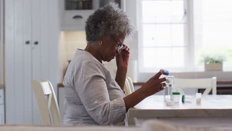 Mujer-Afroamericana-Mayor-Mirando-Un-Contenedor-De-Medicamentos-Vacío-Mientras-Está-Sentada-En-Casa