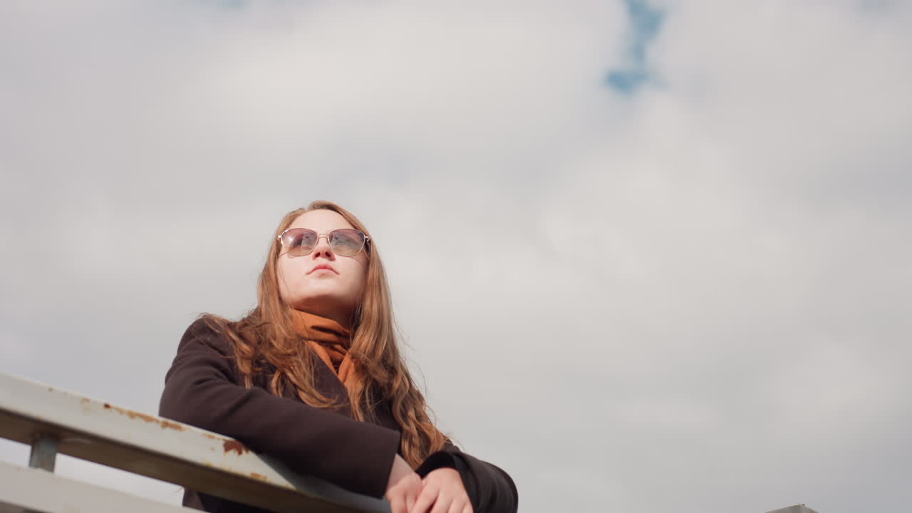 Camera captures calmness of sky reflecting gentle weather condition, creating cool and fresh atmosphere around serene woman leaning on iron bar, showing harmony between light, air, and tranquil mood