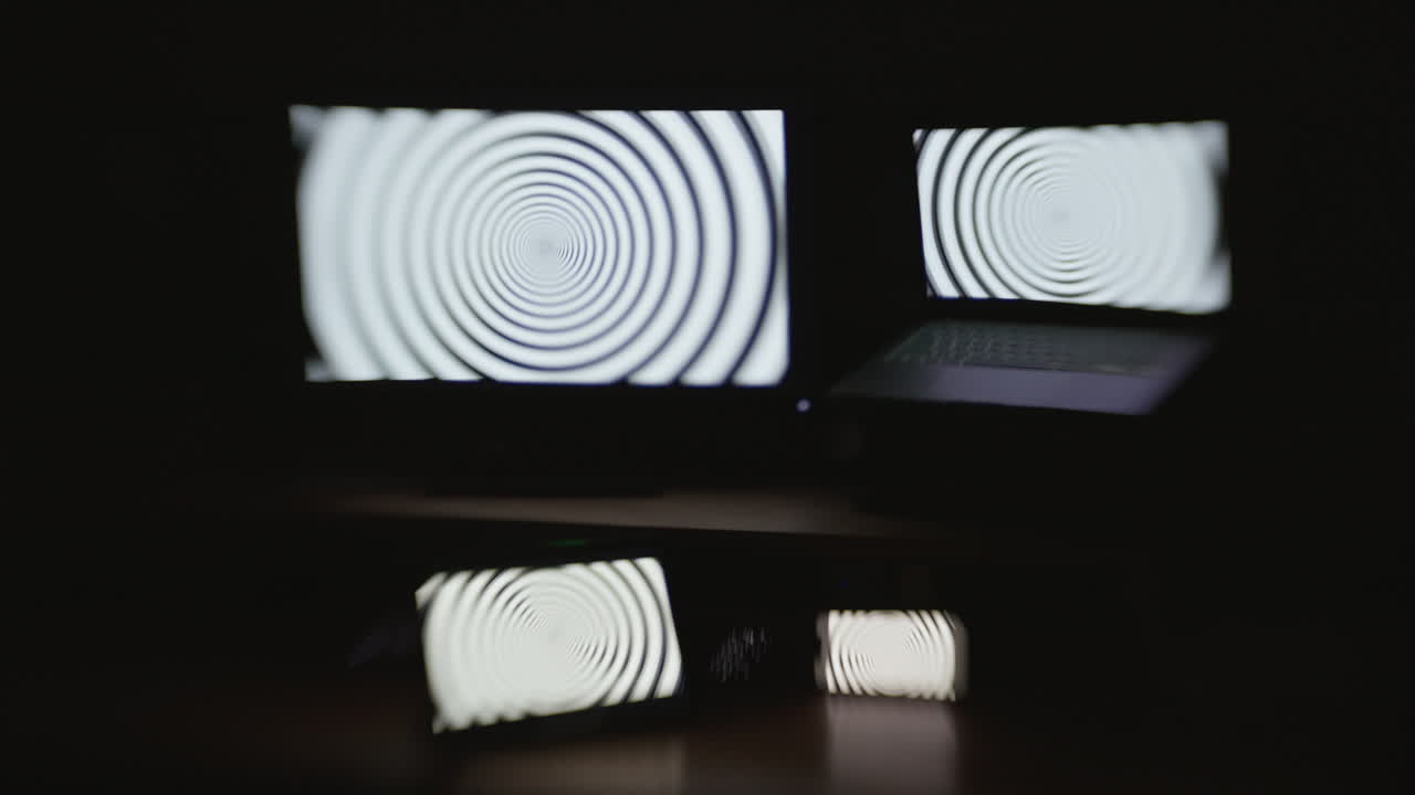 Front angle shot of a hypnotic spiral pattern displayed on four device screens in a dark room with selective focus and a choppy shutter speed effect