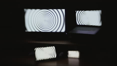 front angle shot of a hypnotic spiral pattern displayed on four device screens in a dark room with selective focus and a choppy shutter speed effect