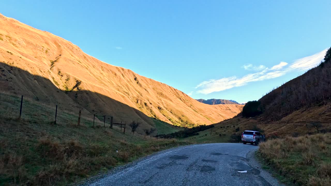 Vehicle travels along gravel road in Glenorchy valley, golden hour sunlight, wide landscape view