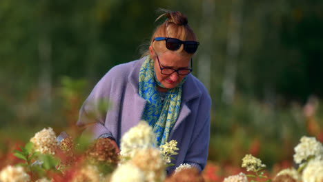 a woman in coat carrying a bag with glasses walks through a flower garden while smelling the floral buds during summer in lithuania