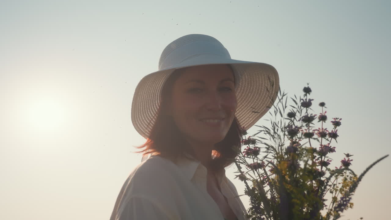 woman holding wildflowers gently scents blossoms as sunlight flares warmly through her white hat, casting peaceful glow across her face in quiet countryside