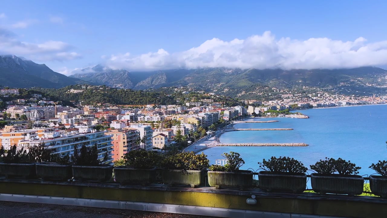 Vista panoramica elevata della costa di Mentone, con edifici colorati, acque calme del Mediterraneo, spiagge, frangiflutti e montagne coperte di nuvole
