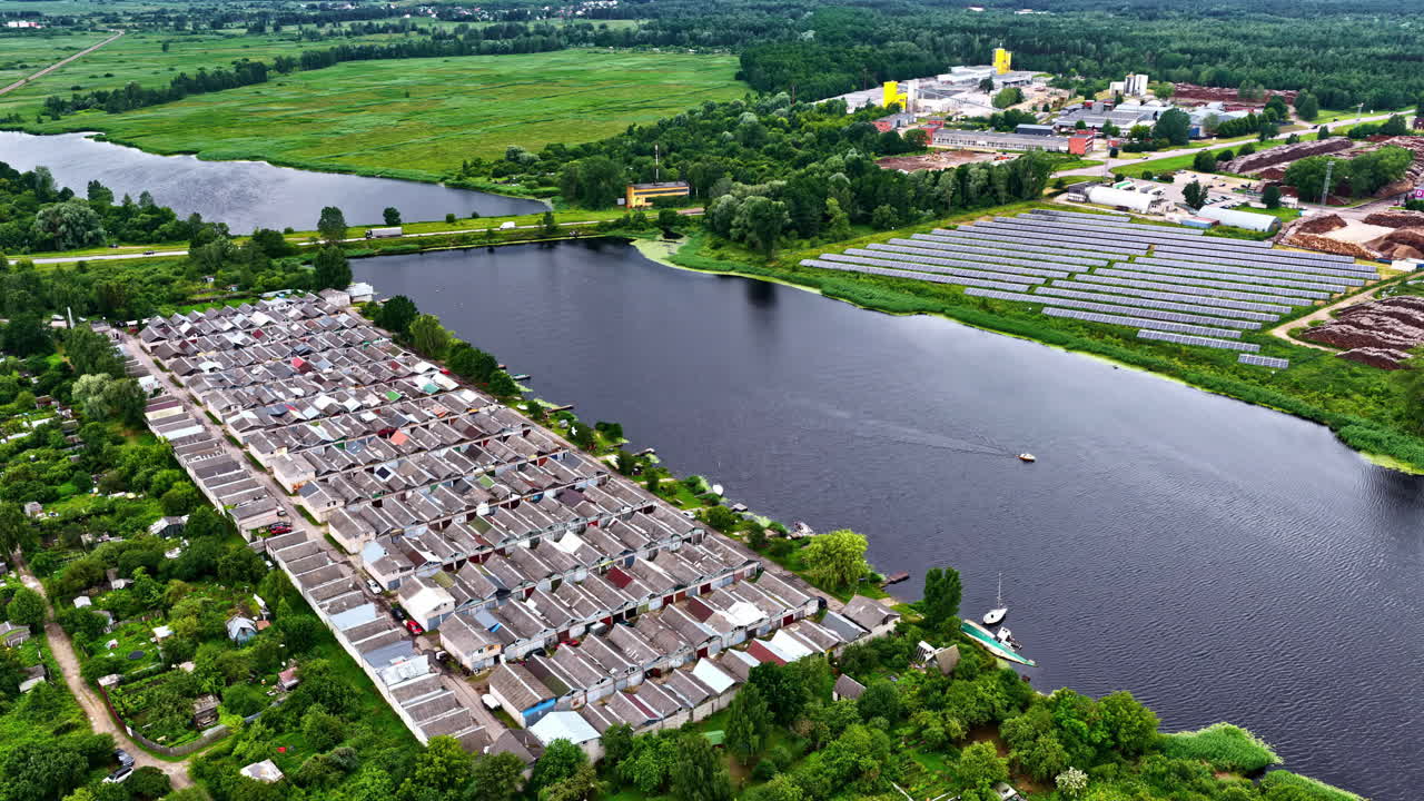 A high aerial view captures a unique Riga landscape with a solar farm generating renewable energy next to a timber factory, bordering the Hapaka Grāvis canal and a dense garage cooperative