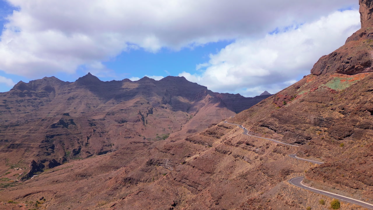 Aerial view of winding mountain road crossing dramatic volcanic landscape in Gran Canaria, Canary Islands, Spain under partly cloudy sky.
