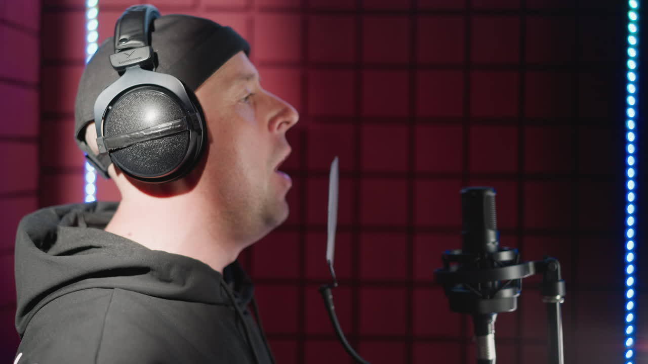 Close-up side shot of a man dressed in black, standing in front of a studio microphone with headphones , passionately singing with visible hand movement in a soundproofed room with red acoustic panels