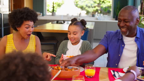 Familia-De-Varias-Generaciones-Sentados-Alrededor-De-La-Mesa-En-Casa-Disfrutando-De-La-Comida-Juntos