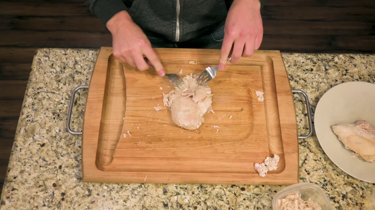 Caucasian male shredding skinless steamed chicken breast with forks on a wooden cutting board