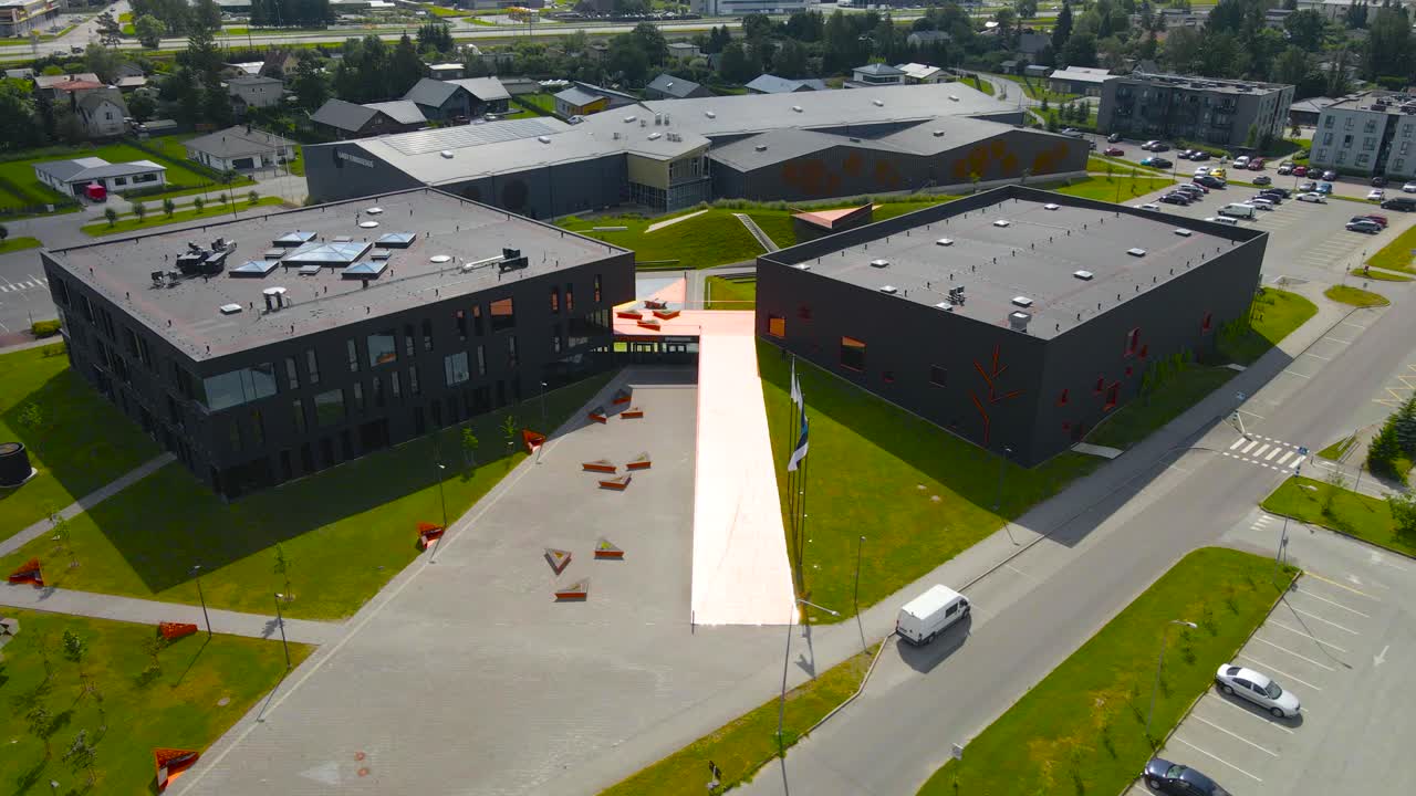 Aerial drone footage orbiting around a black and orange colored modern school building in Saue Laagri during a summer sunny day with green grass and road around while flags wave in front of the school