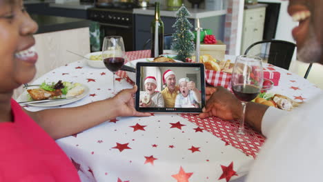 una pareja afroamericana sonriente usando una tableta para una videollamada de navidad con la familia en la pantalla