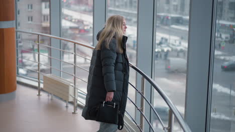 lady standing near iron railing, holding black handbag, gazing out through glass with cityscape and office building in background, people visible moving around in busy urban setting