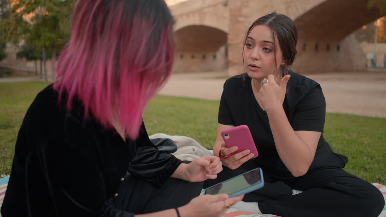 Two women talking and looking at phones in a park