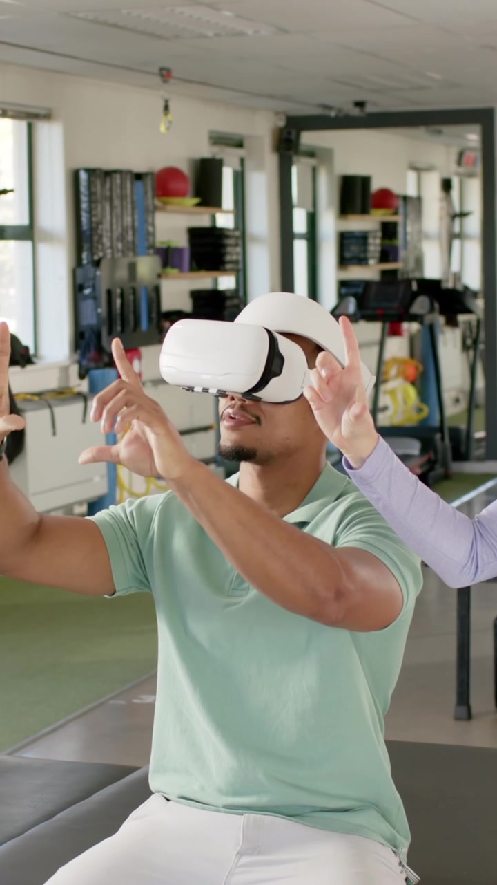 Video verticale: Uomo afroamericano con visore VR bianco, fisioterapista che guida, pizzica, sceglie in riabilitazione