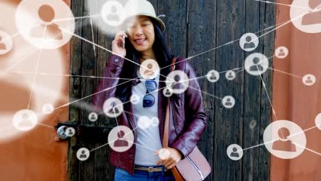 animation of connected profile icons over happy asian young woman talking on cellphone outside door