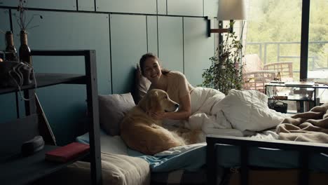 A-brunette-girl-in-a-light-T-shirt-lies-in-bed-and-plays-with-her-big-light-colored-dog-at-home-against-the-background-of-a-turquoise-wall