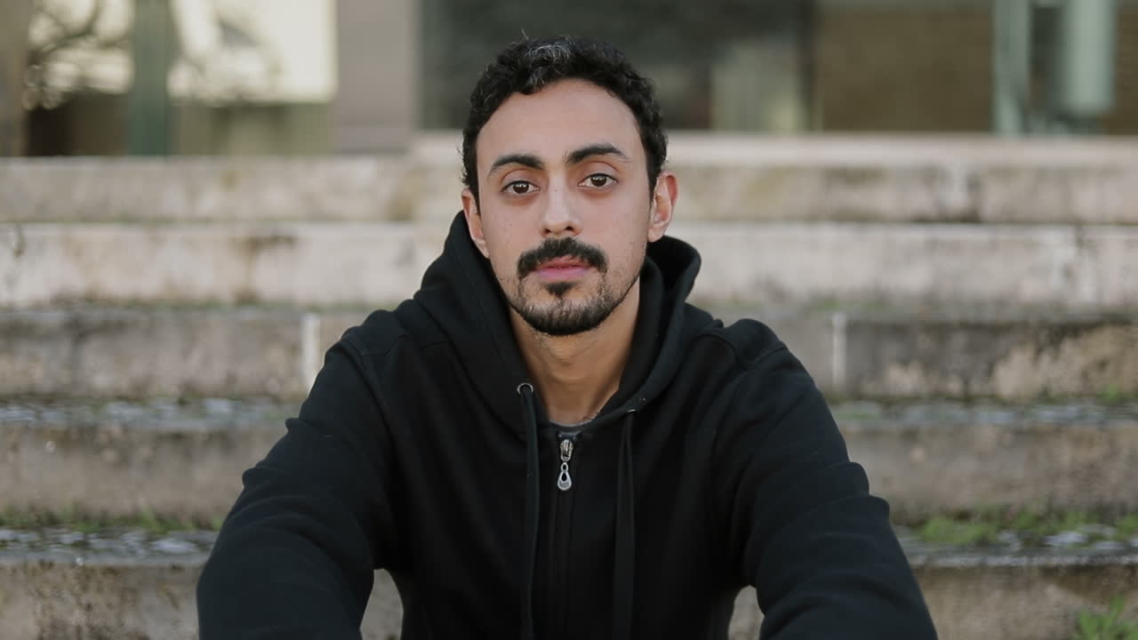 vista frontal de un joven árabe guapo con cabello rizado oscuro y barba con capucha negra sentado en las escaleras afuera
