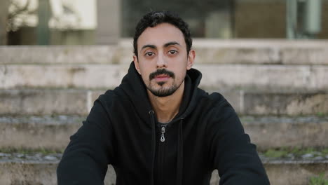 front view of serious young arabic handsome man with dark curly hair and beard in black hoodie sitting on stairs outside