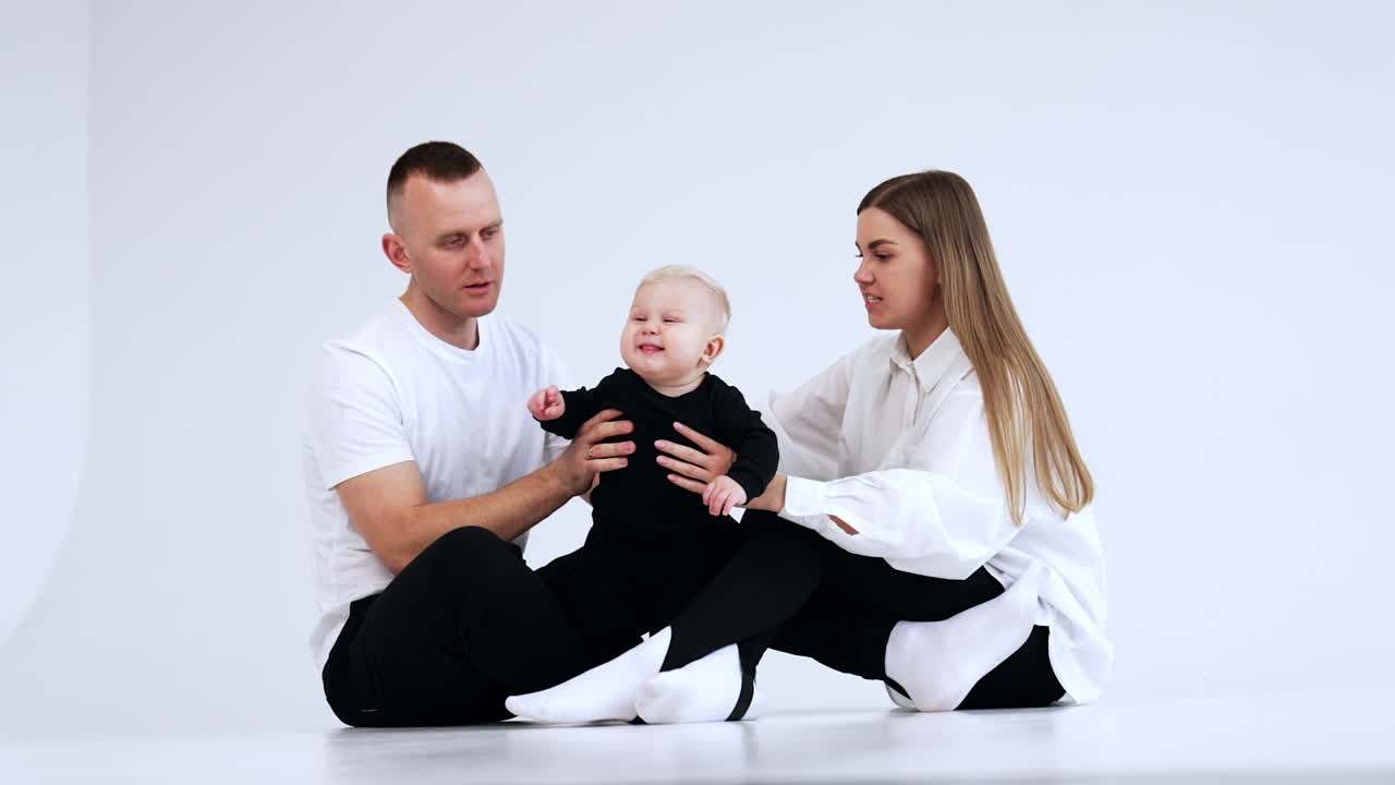 Caucasian parents in white shirts and black trousers sit on the floor. Family portrait with infant baby in white studio.