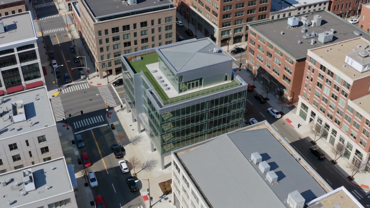 Dron acercándose capturando un edificio de cristal en el centro para mostrar un techo ajardinado y un ático, con coches en movimiento.